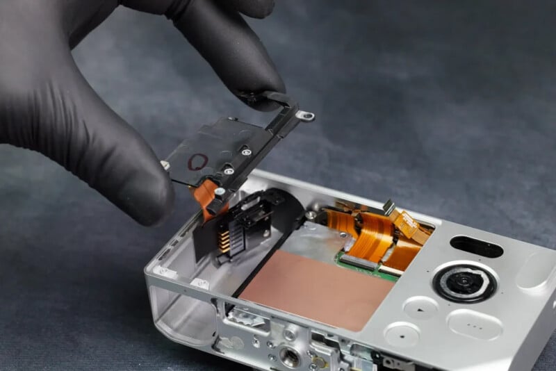 A gloved hand removes an internal component from a partially disassembled electronic device, revealing circuit boards and ribbon cables inside a silver casing.