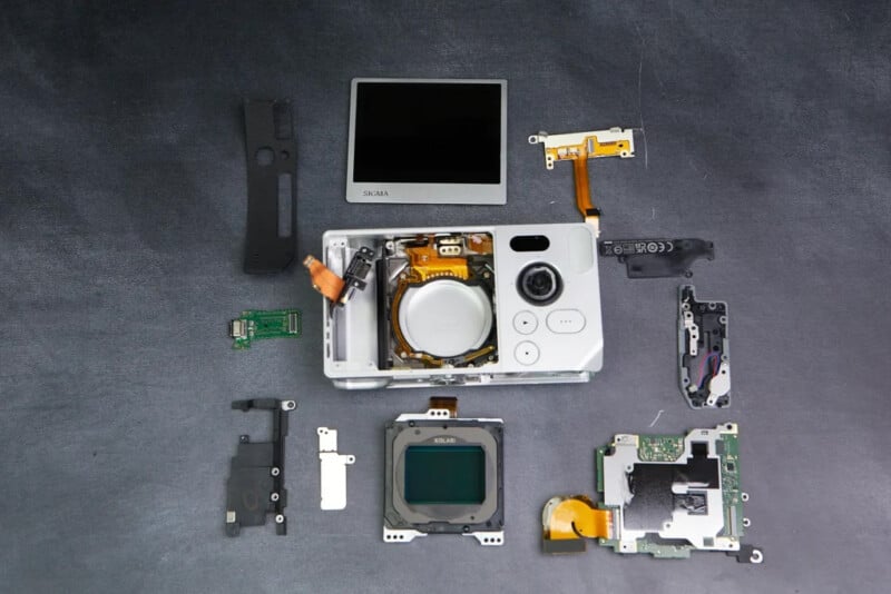 A digital camera is disassembled on a dark surface, with its screen, lens, circuit boards, and various internal components neatly arranged around the camera body.