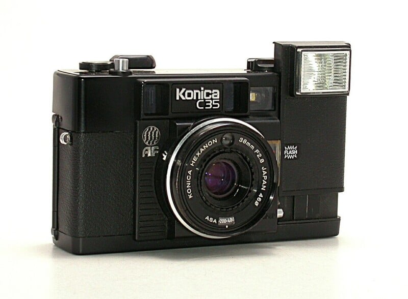 A black Konica C35 film camera with a 38mm lens and built-in flash is positioned on a white surface, facing slightly to the left. The camera has visible dials and textured grip details.
