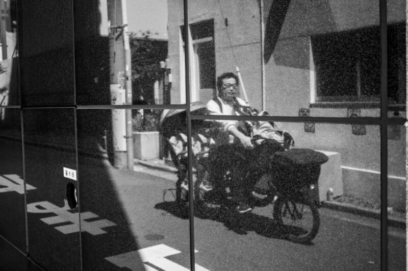 Reflection of a person riding a bicycle with two children, captured on the dark, tiled surface of a building along a city street in black and white.