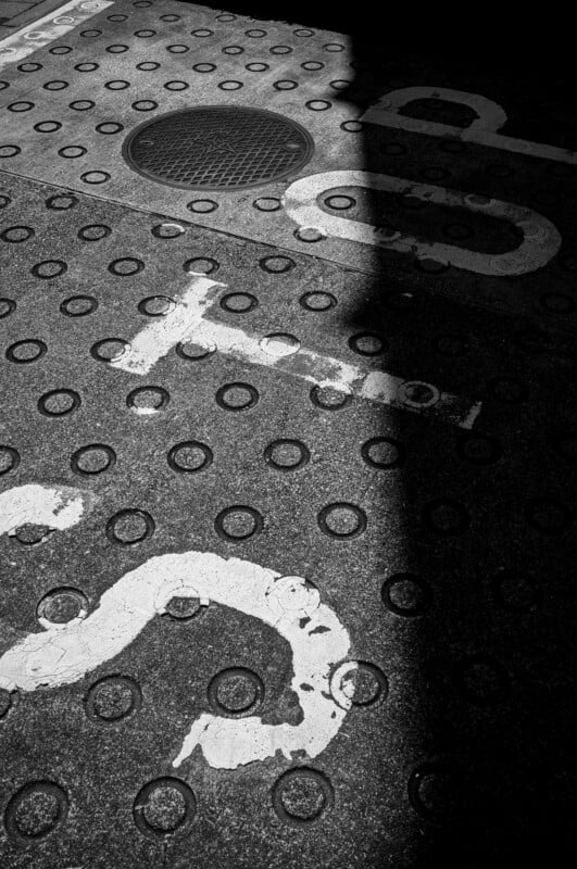 A concrete sidewalk with tactile dots and faded white paint markings, partially in shadow. A circular manhole cover is visible near the top of the image.
