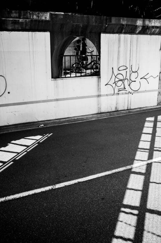 A black-and-white photo of a street underpass with shadows from a railing on the road. A person with a bicycle is visible through a barred window in the wall, which is marked with graffiti.