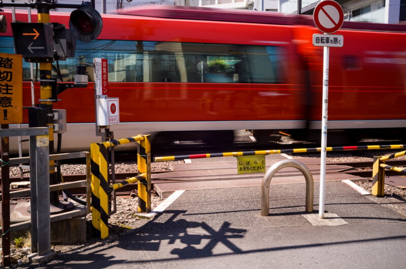 A red train speeds through a railway crossing, with barriers down and warning signs posted. A “no entry” sign is visible, and shadows are cast on the pavement in the foreground.