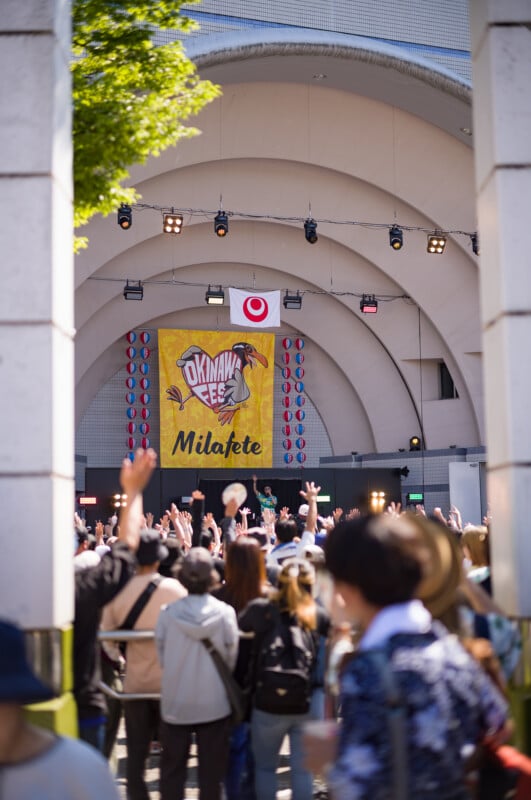 A crowd raises their hands in front of an outdoor stage with a large yellow "Okinawa Milafete" banner and Japanese flag, during a festival under a sunny sky.