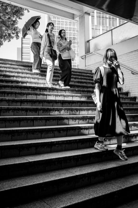 Four women walking on outdoor steps in an urban area; three descend, one holds an umbrella, while another woman in a dress ascends, lit by sunlight. The scene is in black and white.