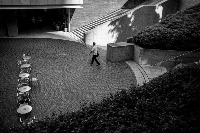 A person walks alone across a paved courtyard with empty metal tables and chairs on one side, stairs and neatly trimmed bushes on the other, in a high-contrast black-and-white urban scene.