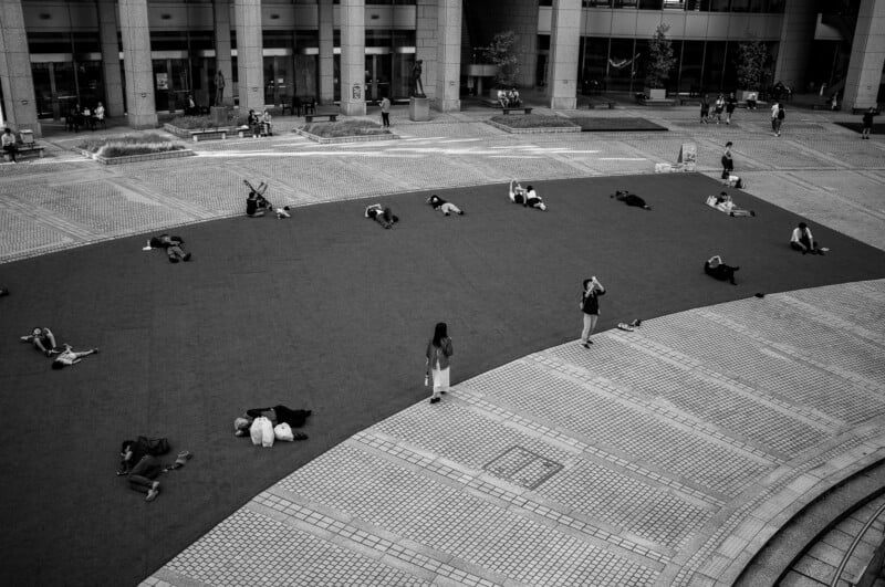 A black and white photo of people lying and sitting on a large, curved patch of grass in an urban plaza, with others walking nearby and tall buildings in the background.