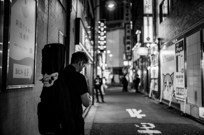 A person with a guitar case on their back stands in a narrow, dimly lit city street at night. Japanese signs and bright storefronts line the alley, and a few people walk in the background. The photo is in black and white.
