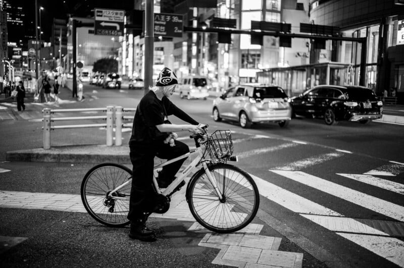 A person wearing dark clothing and a beanie waits on a bicycle at a city crosswalk at night. Bright lights, cars, and buildings fill the busy urban background. The scene is in black and white.