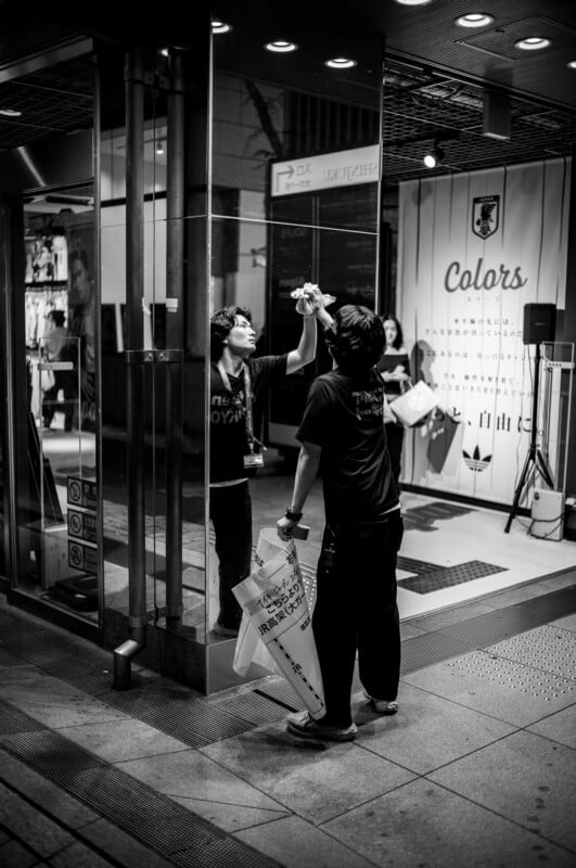 A person cleans a glass storefront at night, holding a cloth and spray bottle, while another stands nearby holding shopping bags. Reflections and store signs are visible in the black-and-white photo.