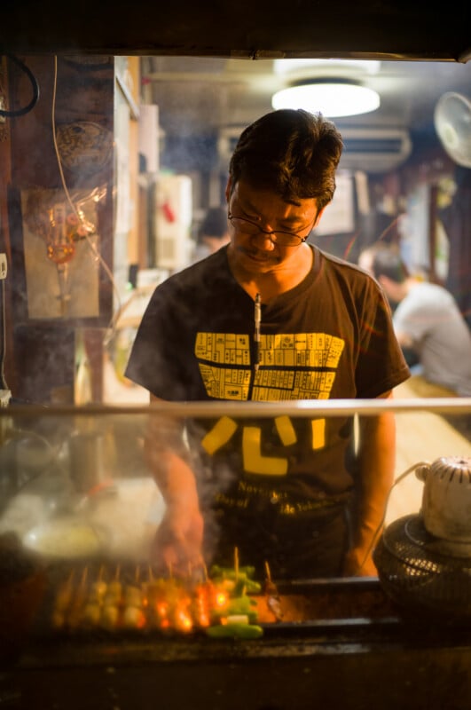 A man cooks skewers over an open grill in a dimly lit food stall, with smoke rising around him. He wears glasses and a black T-shirt with yellow writing, focusing intently on grilling the food.