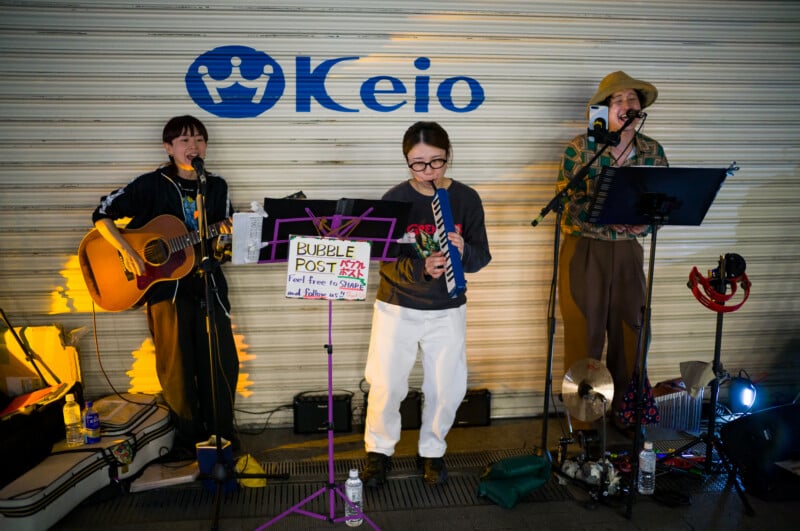 Three street musicians perform in front of a closed shop shutter with a "Keio" sign. One plays guitar and sings, another plays melodica, and the third sings into a mic. A sign reading "Bubble Post" is displayed in front of them.