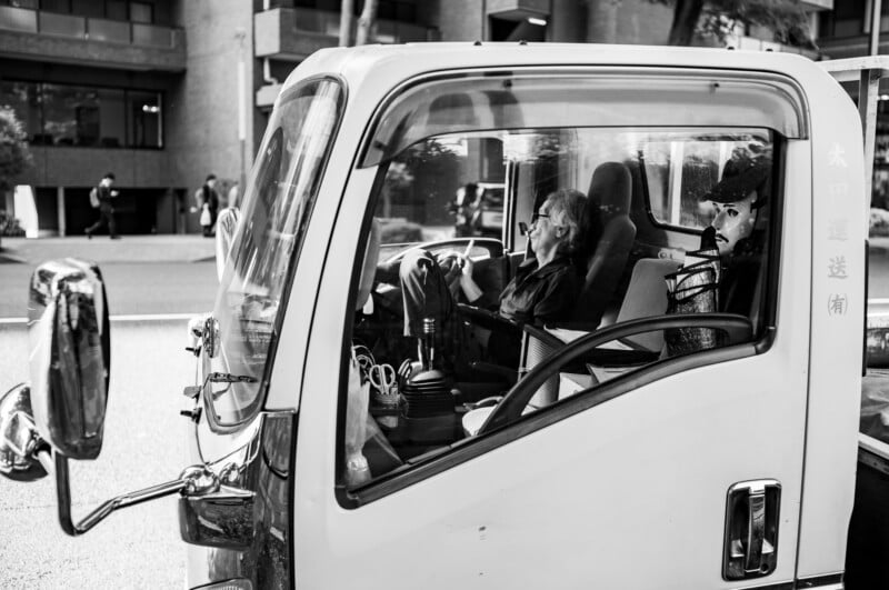 Black and white photo of two men sitting inside a parked truck on a city street. One man is leaning back and relaxing, while the other sits upright. Urban buildings and pedestrians are visible in the background.