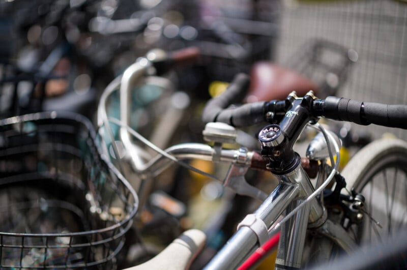 Close-up of several bicycles parked together, focusing on the handlebars and basket of one bike in the foreground. The background is blurred, showing more overlapping bikes, creating a busy, urban scene.