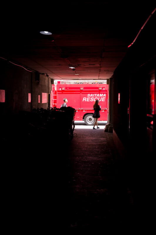A red Saitama Rescue truck is parked outside, seen from a dark tunnel. Two people stand near the truck in the daylight, while bicycles are silhouetted against the light.