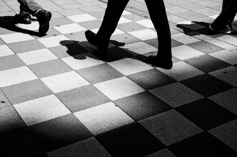 Three people walking on a checkered sidewalk, with their shadows cast on the ground. The image is black and white, focusing on their legs and feet, and strong contrast creates dramatic shadows.