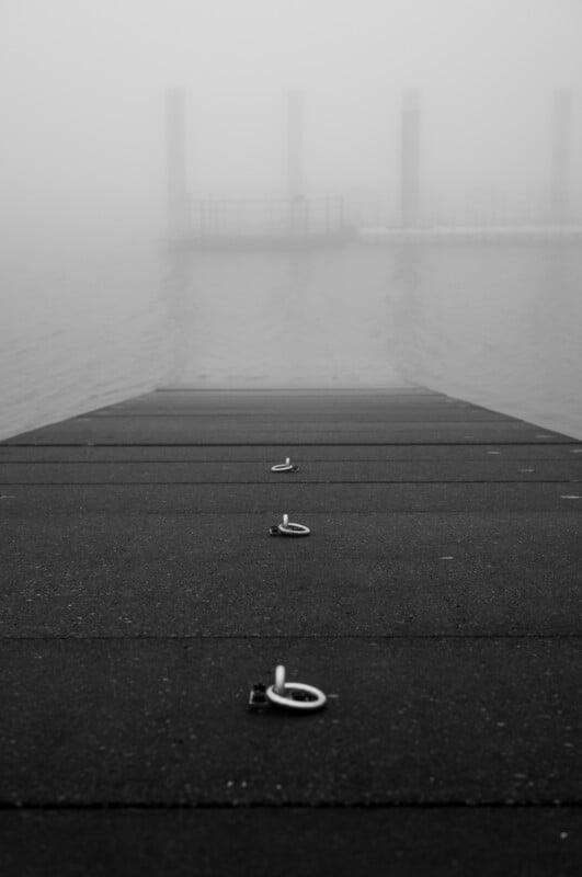 Una escena de niebla que muestra el final de un muelle con ganchos metálicos en la superficie que conducen al agua. Al fondo, los contornos de columnas y plataformas flotantes son apenas visibles en la niebla.