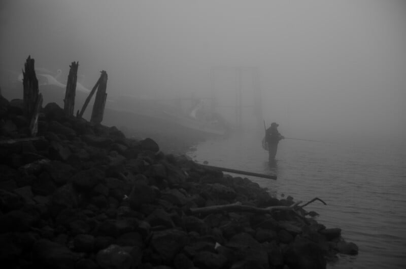 Un hombre pesca con una caña en aguas poco profundas mientras está parado en una costa brumosa, rodeado de rocas y madera flotante, con barcos apenas visibles al fondo.