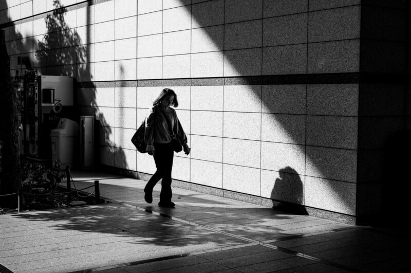 Un hombre camina solo por una acera soleada junto a una pared texturizada que proyecta sombras claras. La escena es en blanco y negro, con zonas claras y oscuras contrastadas.