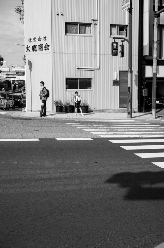 Una fotografía en blanco y negro de un hombre y un niño parados en la acera cerca de un edificio con letras japonesas, esperando que cambie una señal de tráfico. Las sombras y la luz del sol contrastan en la calle.
