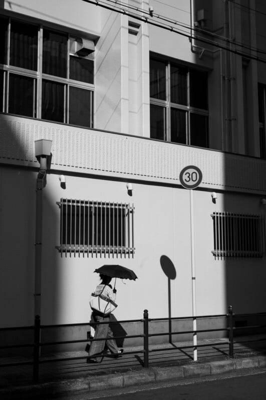 Un hombre caminaba por una calle soleada sosteniendo un paraguas, y su sombra se proyectaba sobre un edificio blanco con ventanas cubiertas por rejas de hierro. "30" Señal de límite de velocidad. La escena es en blanco y negro.