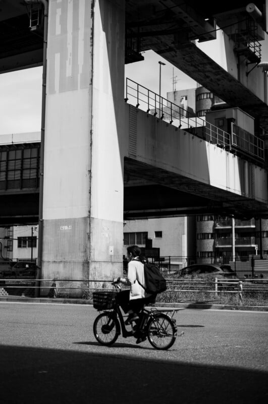 Un hombre anda en bicicleta por una calle de la ciudad bajo una gran estructura elevada de hormigón con edificios altos al fondo. La imagen está en blanco y negro.