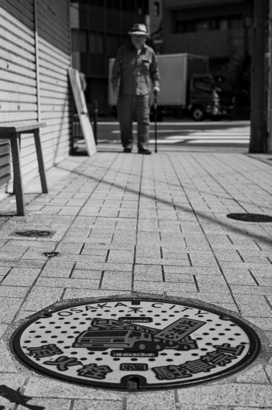 Un hombre con un bastón camina por una acera de la ciudad de Osaka, Japón. En primer plano se muestra una tapa de alcantarilla decorada con la etiqueta "Ciudad de Osaka." Las fotos están en blanco y negro.