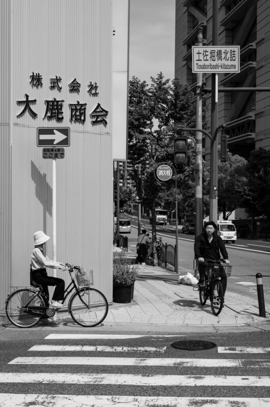 Dos personas circulaban en bicicleta por un paso de peatones de la ciudad. Un ciclista vestía ropa ligera y gorro; el otro vestía ropa oscura. Al fondo se pueden ver símbolos y edificios japoneses. La imagen está en blanco y negro.