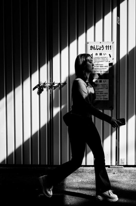 Una mujer camina rápidamente junto a una pared de metal con espectaculares sombras cortadas y un cartel japonés. De lado, sostiene un teléfono móvil y la luz y la sombra crean un fuerte contraste en la escena.