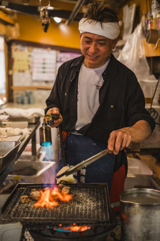Un chef sonriente con una diadema blanca utiliza unas pinzas y un soplete para asar comida sobre un fuego abierto en una cocina acogedora y cálidamente iluminada. Lo rodean varios utensilios e ingredientes de cocina.