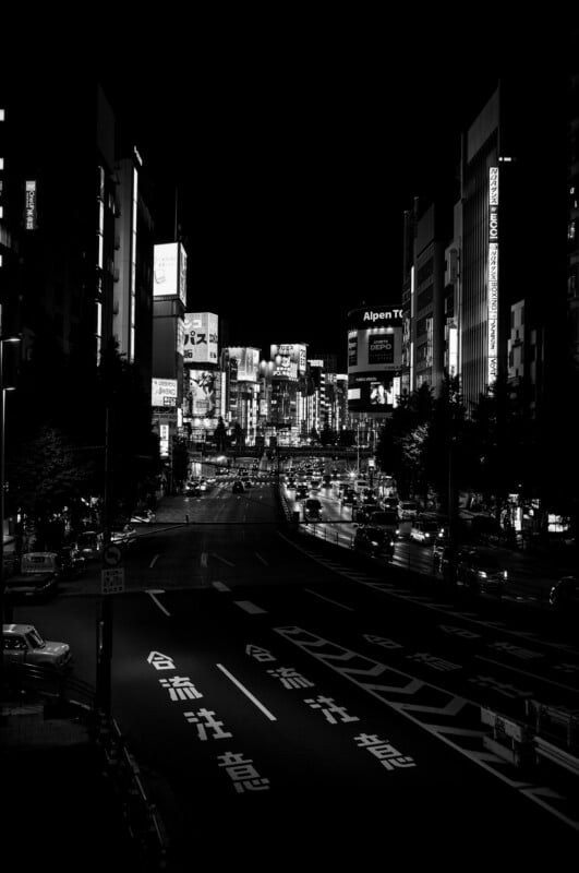 A black and white night view of a busy city street in Japan, illuminated by bright signs and headlights, with Japanese characters painted on the road and tall buildings lining both sides.