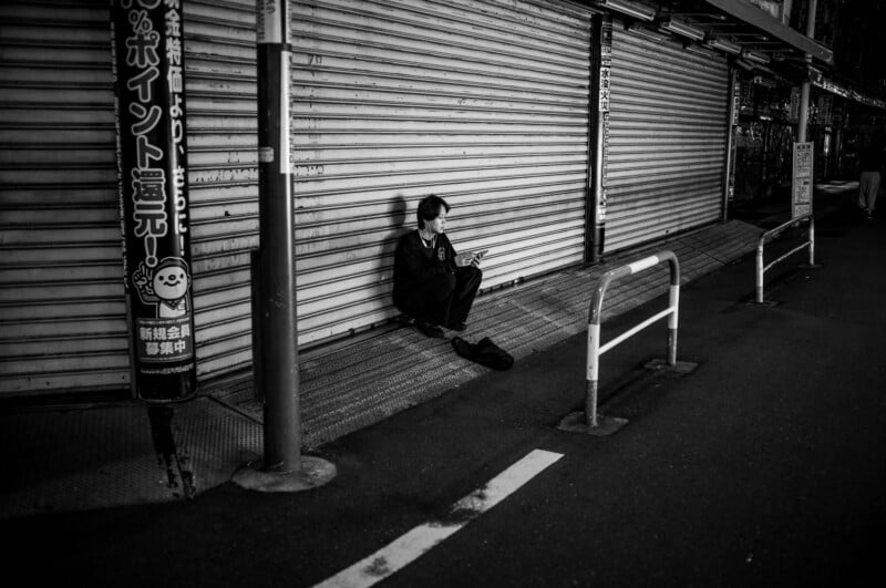 A person sits alone on the sidewalk against a closed shop’s metal shutter at night, looking at their phone. The street is empty and dimly lit, with Japanese signs visible on a pole.
