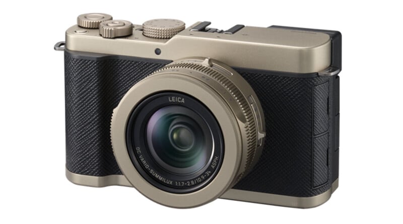 A gold and black Leica digital camera with a large lens, textured grip, and various dials and buttons on the top. The camera is angled slightly to the right.