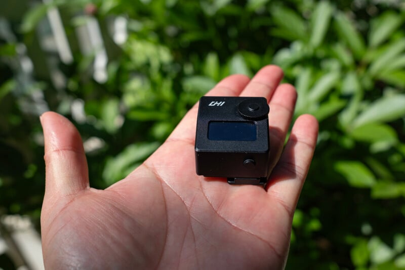 A person holds a small black electronic device labeled "CH1" with a screen and button in their palm. Green leafy plants are blurred in the background.