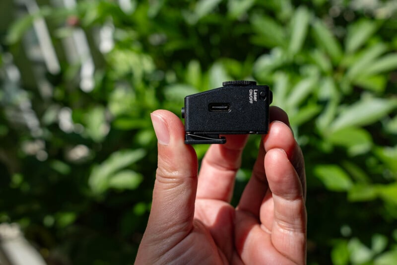 A hand holds a small black camera accessory with a USB-C port and "USB-C CHARGE ONLY" label, against a blurred green leafy background in sunlight.