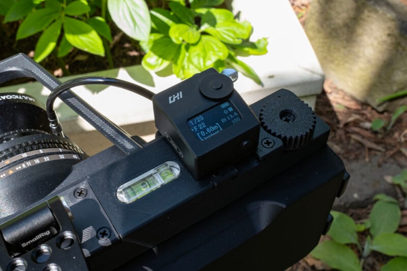 Close-up of a camera with a digital device attached to the hot shoe, showing settings on its screen. The camera is outdoors, surrounded by green plants and sunlight.