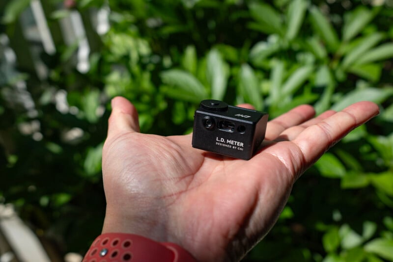 A person holds a small black device labeled "L.D. Meter" in their palm, with green leafy plants in the blurred background. The person's hand is well-lit and they are wearing a red wristband.