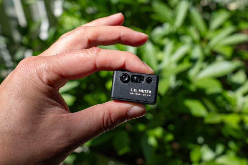 A hand holding a small black device labeled "L.D. METER DESIGNED BY CMJ" outdoors, with green, leafy plants blurred in the background.
