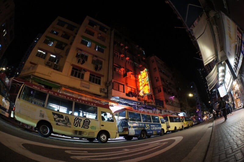 Nighttime street scene with several yellow minibuses parked in front of tall apartment buildings with neon signs and storefronts, captured with a fisheye lens for a distorted perspective.