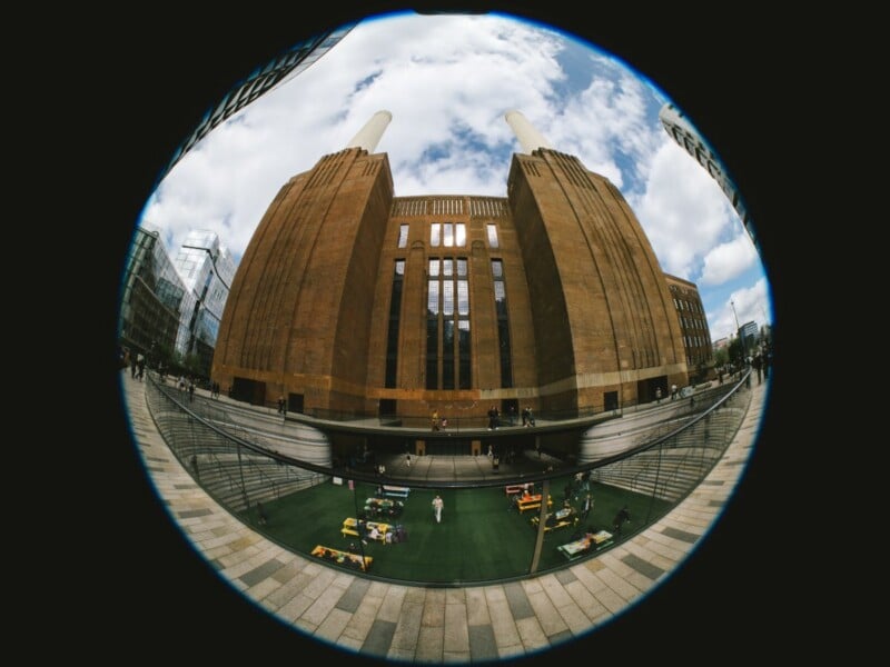 Fisheye lens view of a large, brown brick building with tall chimneys, surrounded by modern glass buildings. People play table tennis in a courtyard below under a partly cloudy sky.