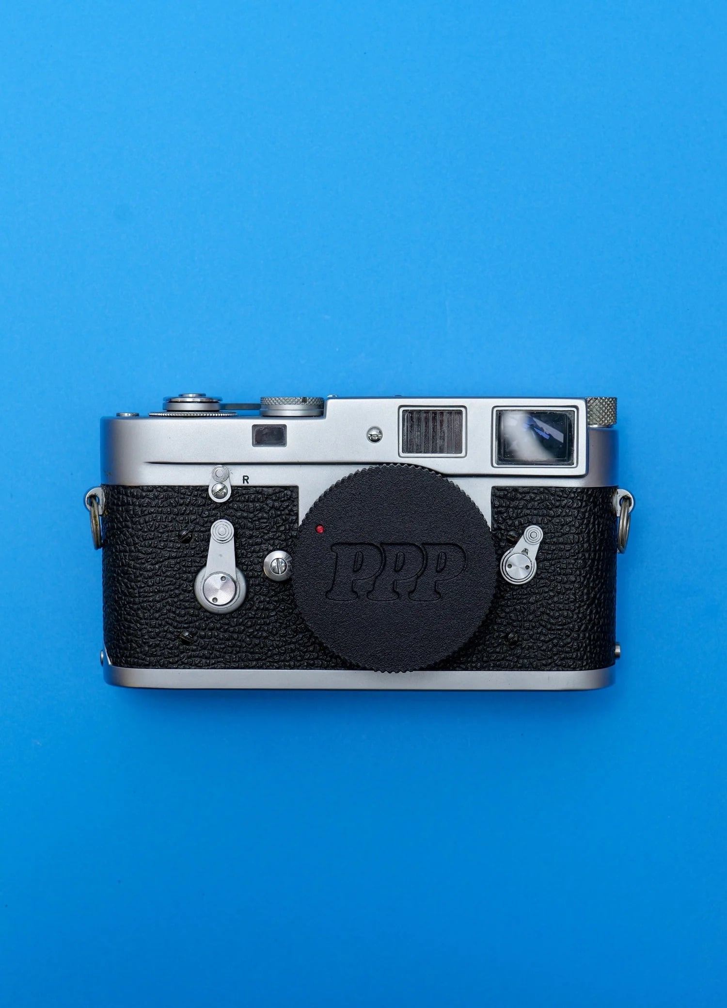 A vintage film camera with a black textured body and silver details is placed on a bright blue background. The camera has a round black lens cap and several buttons and dials visible.