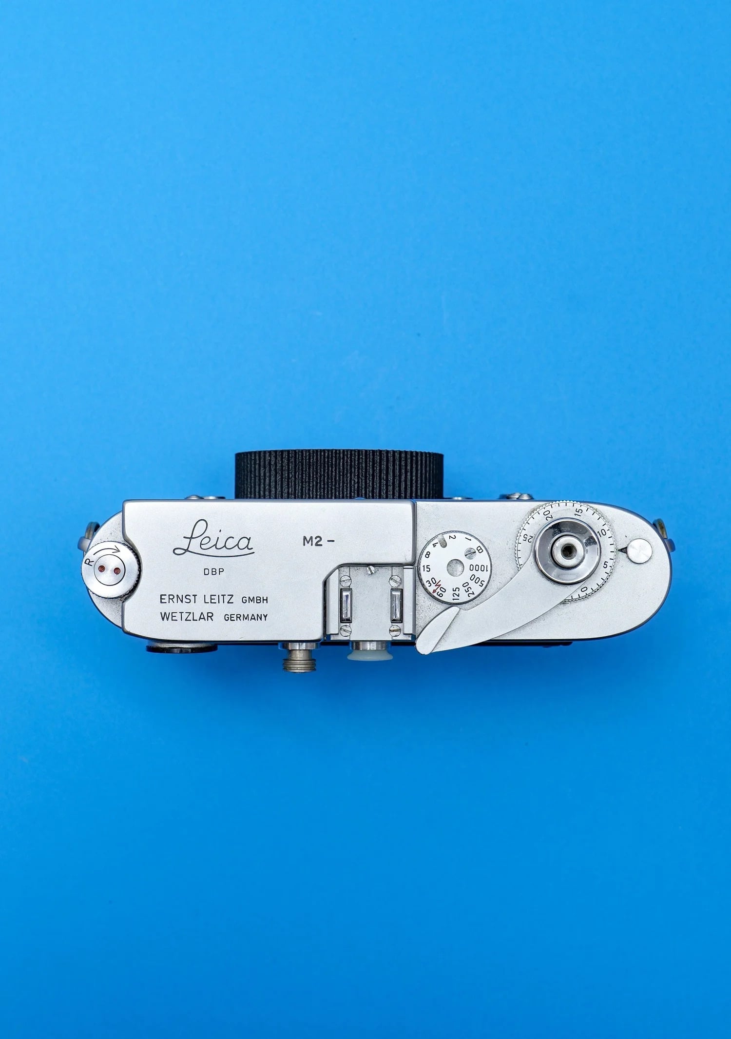Top view of a vintage silver Leica M2 film camera body against a solid blue background, showing the dials, shutter button, and engraved branding.