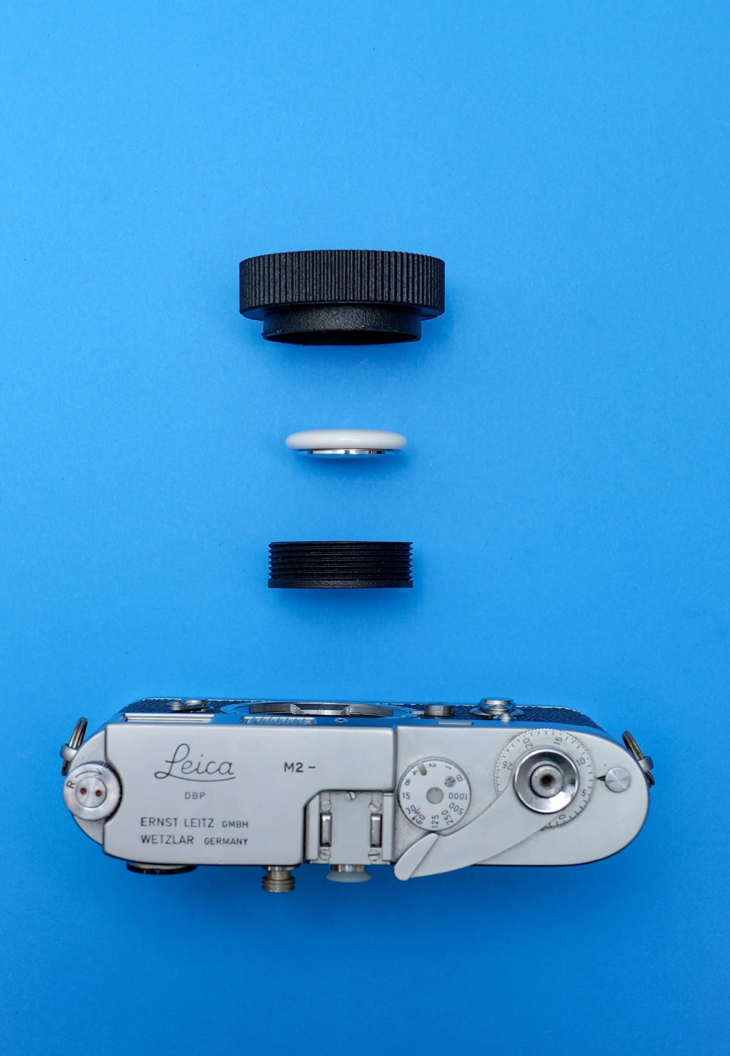 A vintage Leica M2 camera and three lens-related items, including two black lens caps and a white camera part, are arranged neatly on a bright blue background.