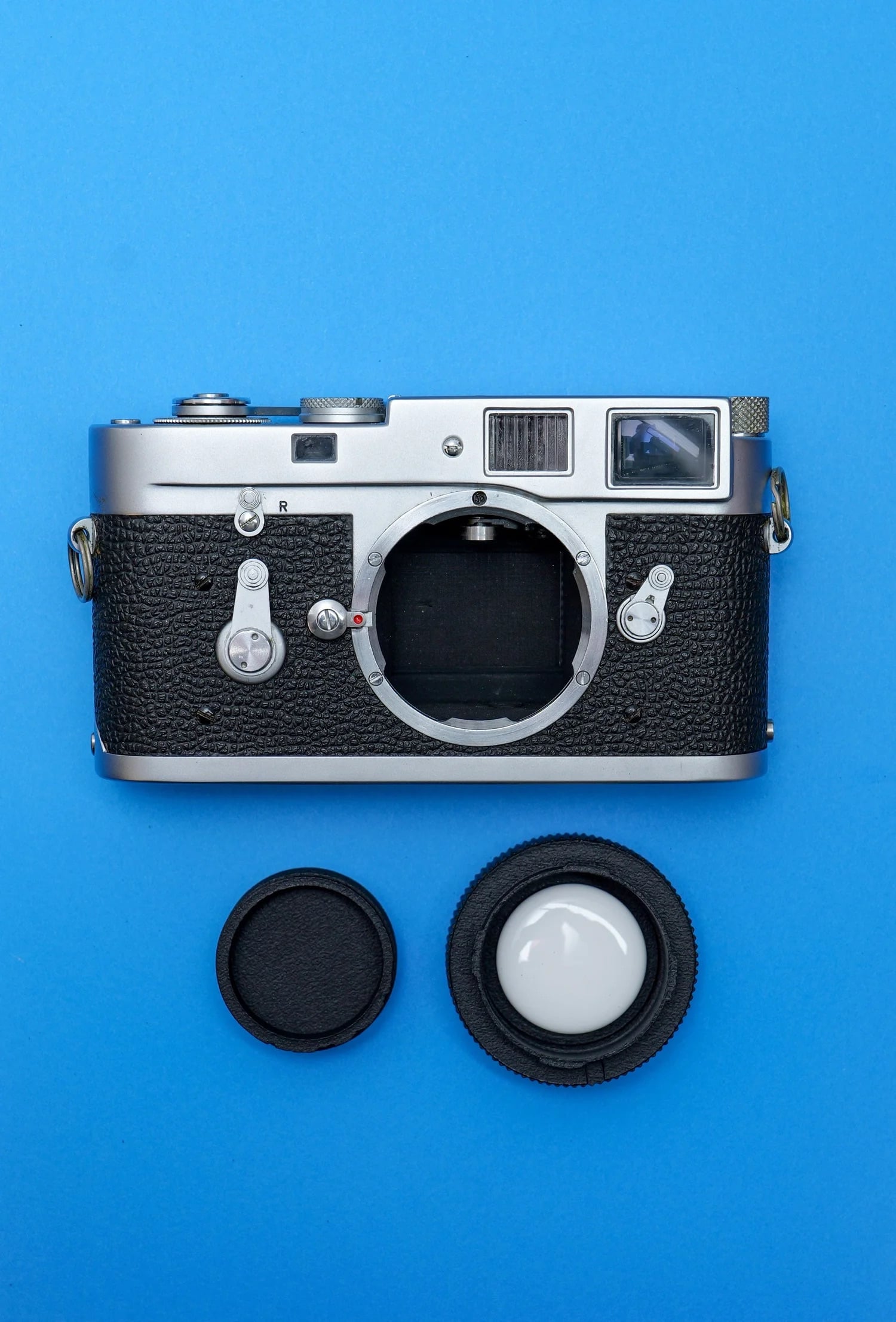 A vintage film camera with no lens attached is placed on a blue background, with two lens caps positioned below it.