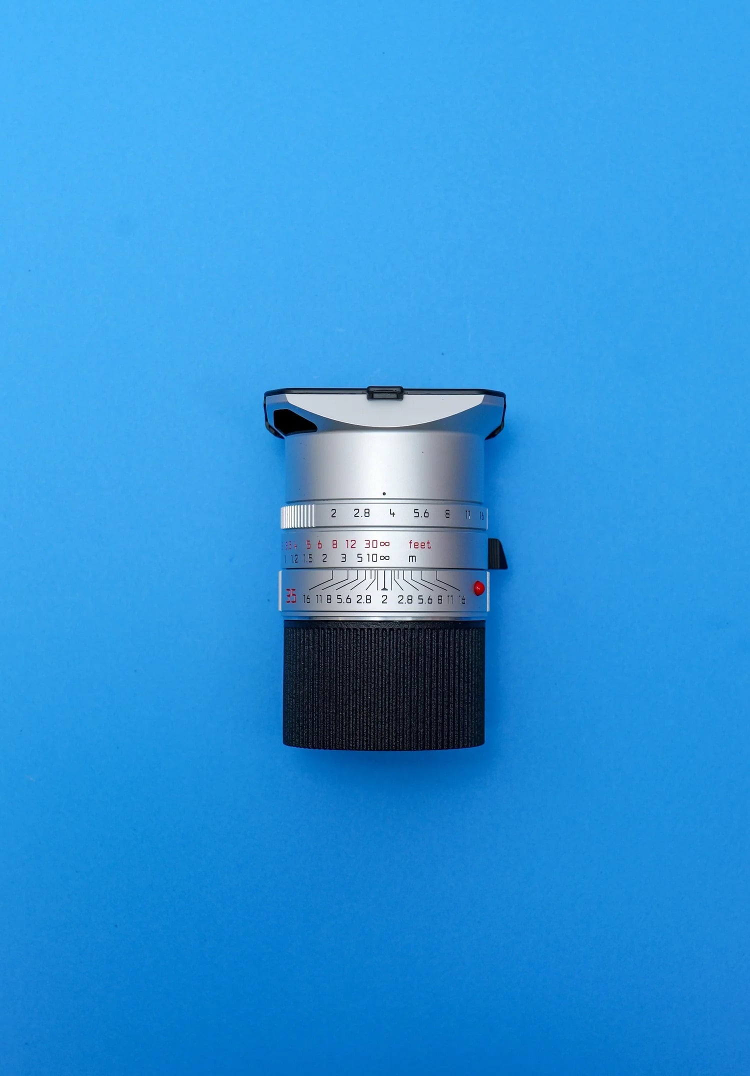 A camera lens with a silver top and black textured bottom is centered on a solid blue background. The focus and aperture markings are visible on the lens barrel.