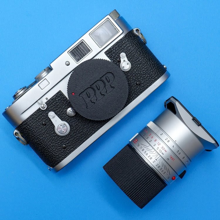 A vintage silver and black camera with a black lens cap marked "PPP" rests on a blue surface next to a detached camera lens with a silver and black finish featuring numbered settings.