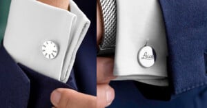 Close-up of a person wearing a navy suit and white shirt, showing silver cufflinks; one has engraved camera settings, the other has “Leica” written in script.