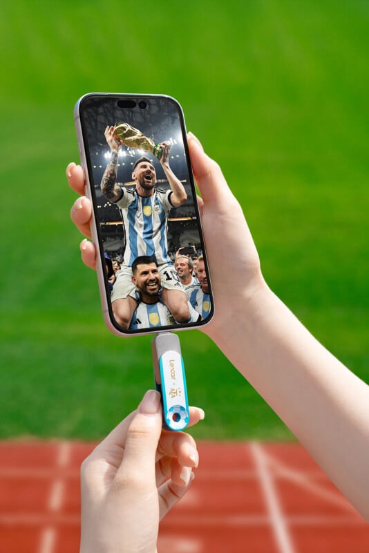 A person holds a smartphone displaying a soccer victory celebration and connects it to a blue USB flash drive shaped like a soccer ball, with a blurry sports field in the background.