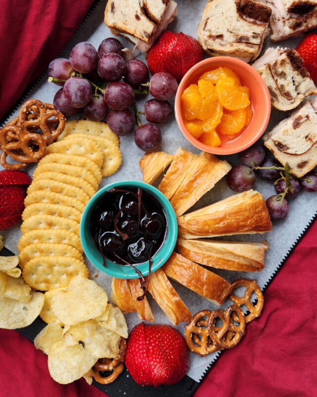 A charcuterie board with crackers, potato chips, pretzels, grapes, strawberries, cinnamon swirl sandwiches, orange slices in a bowl, pastry slices, and a bowl of dark cherries, set on a red cloth background.