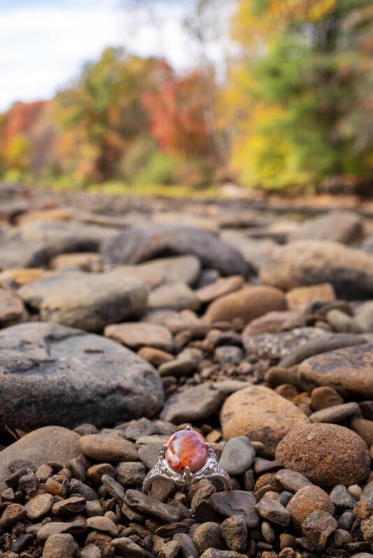 A gemstone ring with an orange-red stone rests on a bed of river rocks, with blurred autumn trees and foliage in the background.